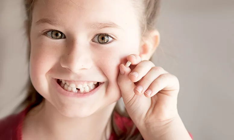 Kleines lachendes Mädchen mit Zahnlücke und einem Milchzahn in der Hand